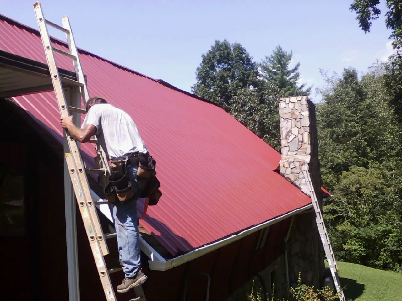 Roofer on a ladder beside a red metal roof and stone chimney — Soffit & Fascia Repair by Roofing Brothers in Saylorsburg